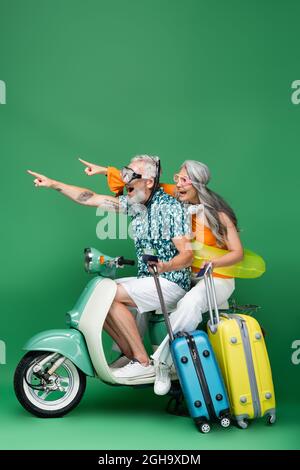 un couple d'âge moyen, émerveillé et multiethnique dans des lunettes de protection pointant avec les doigts et tenant un passeport tout en voyageant sur un scooter à moteur vert Banque D'Images