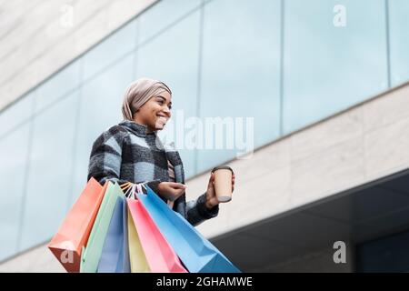 Joyeuse jolie femme noire musulmane dans le hijab et manteau avec des sacs d'achats et de café à emporter, en appréciant le shopping Banque D'Images