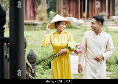 Belle femme et homme vietnamiens souriant dans les robes ao dai tenant des fleurs de lotus en entrant dans le vieux bâtiment Banque D'Images