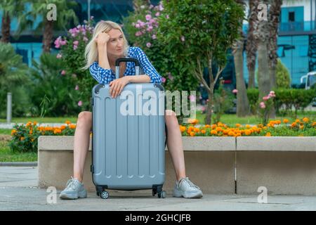 Une jeune fille blonde pensive est assise sur un banc de parc un jour d'été, penchée ses mains sur une valise grise debout entre ses jambes Banque D'Images