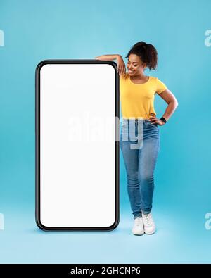 Joyeuse Afro femme dans une tenue décontractée penchée sur un énorme téléphone portable avec écran blanc vide sur fond bleu, maquette Banque D'Images