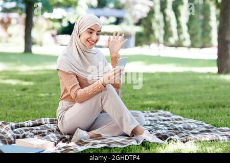 Une jeune femme musulmane arabe heureuse dans le hijab appelle sur un smartphone, réunion vidéo dans le parc vert Banque D'Images