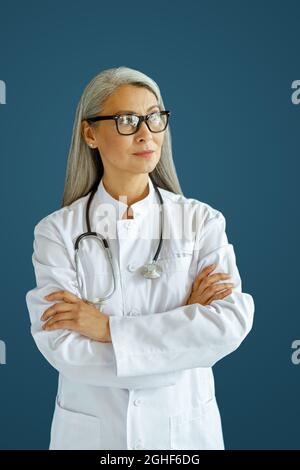 Femme expérimentée mature médecin avec des lunettes et des bras croisés se tient sur fond bleu Banque D'Images