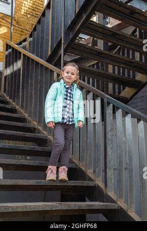 Petite fille blonde caucasienne dans un jeune tendance chaud vêtements de dessus de printemps se tient sur l'escalier de fer vintage regardant la caméra. Très beau Banque D'Images