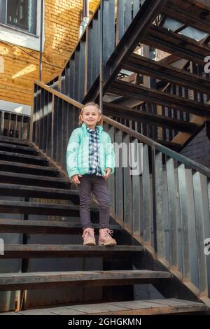 Petite fille blonde caucasienne dans un jeune tendance chaud vêtements de dessus de printemps se tient sur l'escalier de fer vintage regardant la caméra. Très beau Banque D'Images