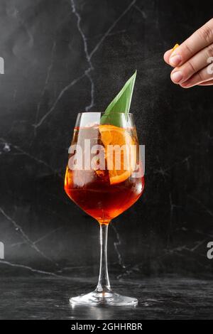 Verre de glace cocktail apéritif servi dans un verre de vin, décoré de tranches de feuilles d'orange et de basilic, placé sur un noir Banque D'Images