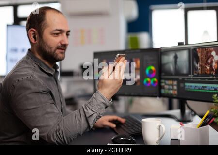 Créateur multimédia artiste parlant sur vidéocall avec le client tenant le smartphone éditant des séquences vidéo assis dans l'agence de production de créativité. Vidéaste utilisant un logiciel de post-production Banque D'Images