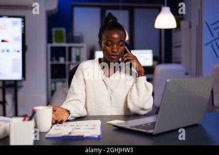 Frustrée exténuée femme d'affaires afro-américaine stressante discutant de stratégie financière avec collegue au téléphone surtravaillant dans le bureau de démarrage. Responsable fatigué analysant les statistiques annuelles de l'entreprise Banque D'Images