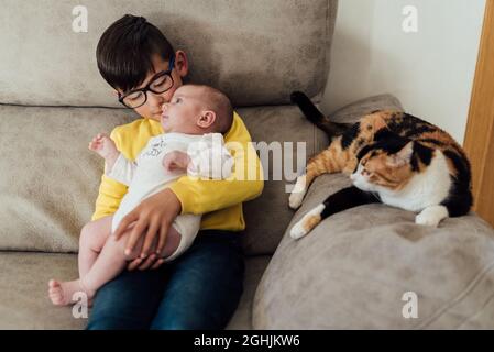 Garçon tenant sa petite sœur dans les bras tout en étant assis sur un canapé avec son chat sur le côté. Banque D'Images