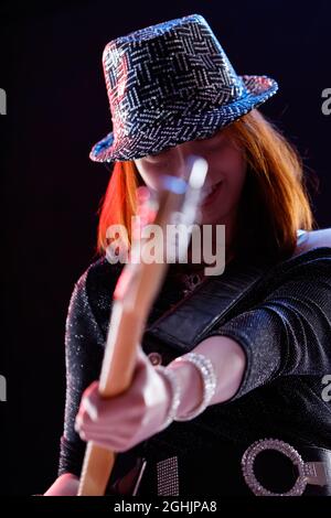 Jeune musicienne tendance et élégante portant un chapeau jouant de la guitare sur scène en souriant alors qu'elle regarde la caméra sur fond sombre Banque D'Images