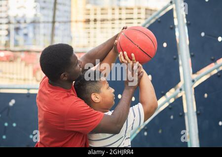 Père afro-américain en t-shirt orange ajustant la position de la main du fils tout en lui enseignant de lancer le basket-ball dans le cerceau Banque D'Images