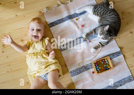 Vue du haut mignonne bébé fille en robe ayant amusant couché sur le sol avec chat jouant des perles amovibles en bois Banque D'Images