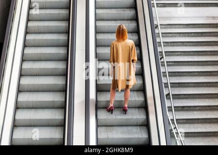 Vue arrière du corps de la femme méconnue dans une robe élégante et des talons hauts debout seuls sur les escaliers de l'escalier roulant souterrain qui se déplace vers le haut Banque D'Images