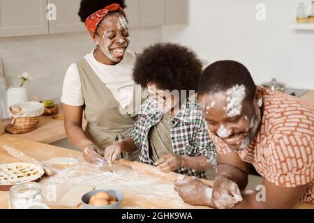 Parents joyeux et leur petit fils s'amusant tout en cuisinant une pâtisserie maison près d'une table de cuisine Banque D'Images