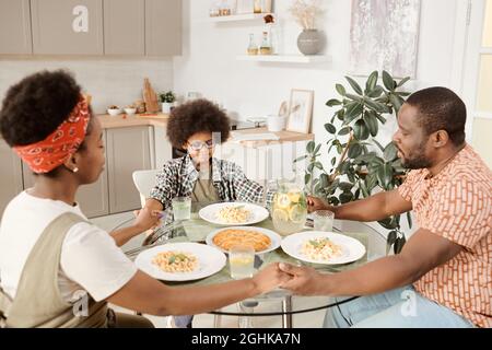 Jeune famille africaine de trois priant avant le dîner par table servie avec leurs yeux fermés Banque D'Images