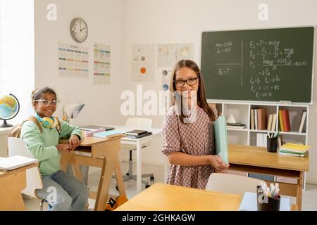 Deux écolières interculturelles vous regardent en classe contre le tableau noir et le lieu de travail de l'enseignant Banque D'Images