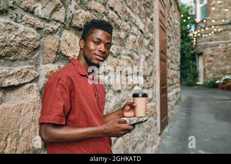 Joyeux jeune homme africain dans les vêtements décontractés défilant dans le smartphone et de boire tout en se tenant par le mur de pierre Banque D'Images