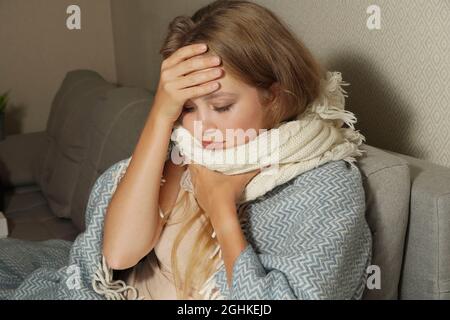 La femme éternue dans un mouchoir blanc. Jeune femme blonde enveloppée dans une couverture chaude, éternuant, a la grippe. Banque D'Images