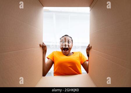 Jeune Afro dame regardant dans la boîte à colis, criant OMG dans l'excitation, obtenir la commande en ligne à l'intérieur, vue de dessous Banque D'Images