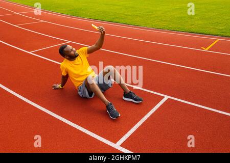 Homme sportif afro-américain en t-shirt jaune assis sur le circuit du stade, en utilisant un smartphone pour prendre un selfie, souriant. L'entraîneur homme est heureux Banque D'Images