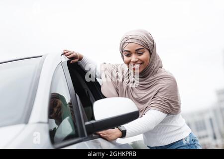 Satisfaite mignonne islamique africaine client américain dans hijab touchant la nouvelle voiture à l'extérieur, appréciant l'achat Banque D'Images