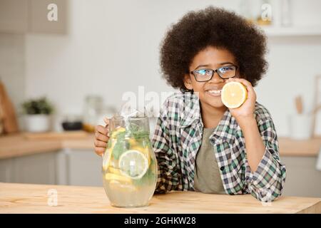 Adorable petit garçon tenant une carafe avec une limonade et une moitié de citron frais tout en se tenant près d'une table de cuisine Banque D'Images