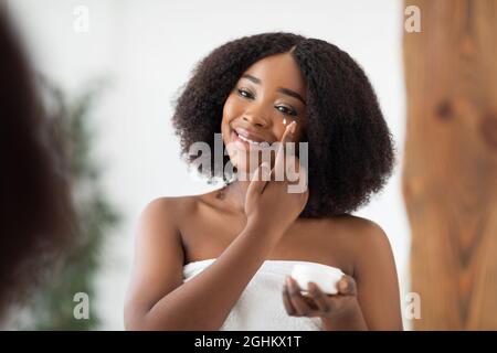 Jeune femme afro attrayante appliquant sous la crème des yeux après le bain près du miroir à la maison. Concept de bien-être domestique Banque D'Images