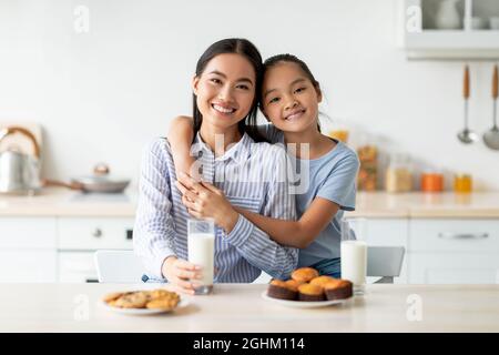 Une mère coréenne aimante et sa fille savourant des petits gâteaux frais faits maison, buvant du lait, assis à l'intérieur de la cuisine Banque D'Images