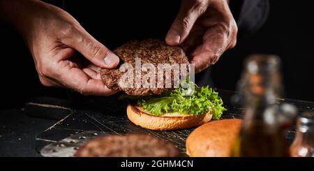 Chef cuisinier qui prépare un hamburger avec une succulente patty de bœuf grillé maison en la plaçant sur une laitue fraîche sur un pain grillé en gros plan sur les mains Banque D'Images