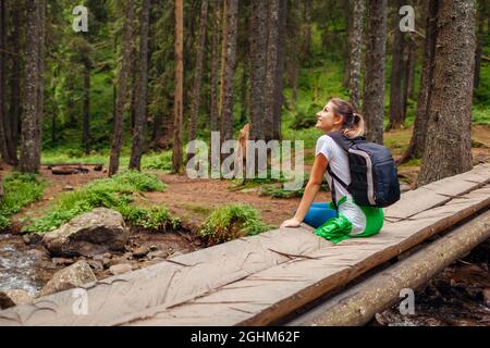 Randonneur voyageur avec sac à dos assis sur le pont de l'autre côté de la rivière dans la forêt carpathienne reposant. Voyage à l'été Ukraine Banque D'Images