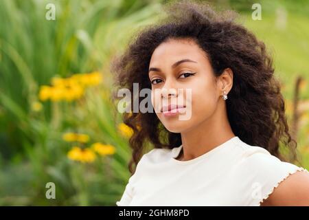 Mixte race noire femme regardant l'appareil photo sourire. Femme afro-américaine avec coiffure afro dans le parc de la ville. Femme à la peau sombre sur fond vert. Mise au point douce. Style tendance minimaliste Banque D'Images