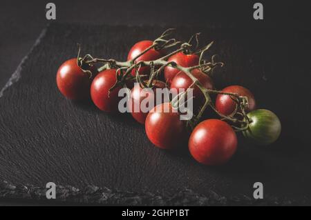 Un bouquet de tomates cerises fraîches sur une branche se trouve sur un plateau de service noir sur fond sombre Banque D'Images