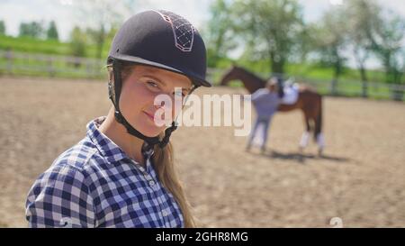 une jeune fille avec un casque sur sa tête regarde une fille floue debout à côté d'un cheval brun et regarde la caméra. Photo de haute qualité Banque D'Images