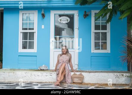 Jolie jeune femme caucasienne avec des cheveux blonds assis près d'un café confortable avec des murs bleus et souriant avec les dents. Photo d'été. Sac en paille tissée Banque D'Images