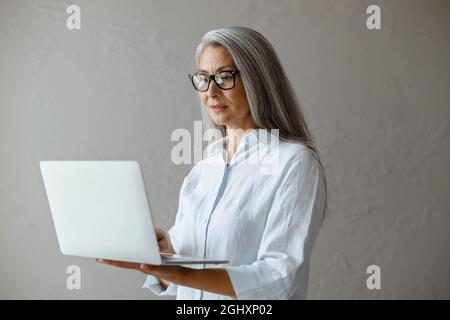 Femme asiatique mature en blouse blanche travaille sur ordinateur portable debout près du mur en pierre grise Banque D'Images