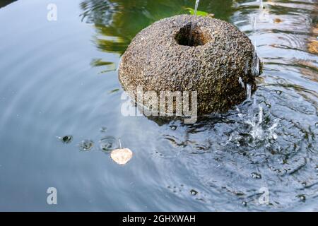 Détail d'un petit étang avec une fontaine en pierre dans laquelle vous pouvez voir les vagues de l'eau sur la surface et une feuille d'automne flottant dessus. Copier l'espace. Banque D'Images