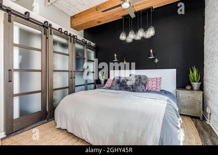 Une chambre dans un appartement loft avec un plafond en bois exposé, des portes coulissantes en verre vers un placard, un mur d'appoint noir, et une lumière de verre de fantaisie suspendue au-dessus. Banque D'Images