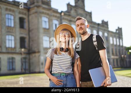 Joyeux lycéens couple souriant. Deux étudiants joyeux, hommes et femmes, avec des sacs à dos debout souriant sur le campus de l'université tenant un ordinateur portable. Photo de haute qualité Banque D'Images