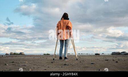 Une femme sur des béquilles marche le long de la rive du lac. Plage de sable sur fond de lac calme et de nuages à l'horizon Banque D'Images