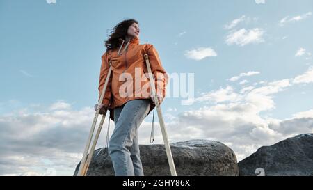 Une femme sur des béquilles marche le long de la rive du lac. Plage de sable sur fond de lac calme et de nuages à l'horizon Banque D'Images