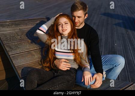 un jeune couple amoureux, un homme et une femme, embrassent un banc dans l'aire de loisirs. Une belle fille aux cheveux rouges dans un chandail et un Jean, un jeune homme attrayant aux cheveux blonds. Le couple heureux. Banque D'Images