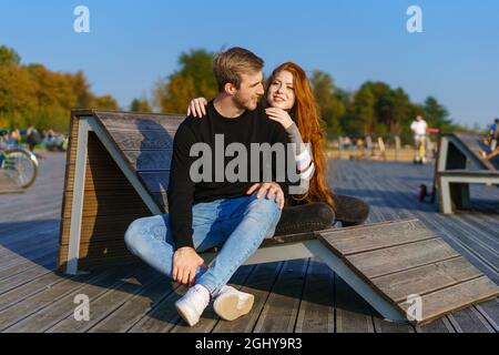 un jeune couple amoureux, un homme et une femme, embrassent un banc dans l'aire de loisirs. Une belle fille aux cheveux rouges dans un chandail et un Jean, un jeune homme attrayant aux cheveux blonds. Le couple heureux. Banque D'Images