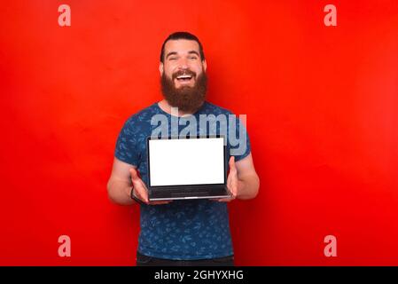 Grand homme barbu souriant tient un ordinateur portable ouvert avec écran blanc sur fond rouge. Banque D'Images