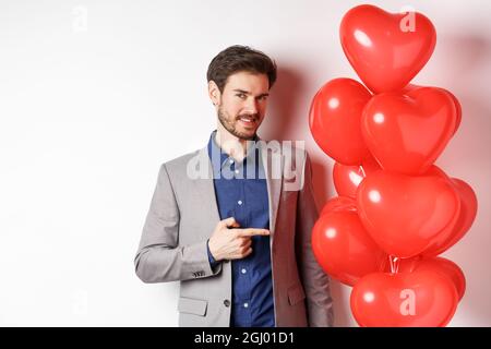 Journée des amoureux. Charmant jeune homme à la barbe, portant un costume de fantaisie, pointant du doigt vers le cœur surprise ballon pour la Saint Valentin, debout sur blanc Banque D'Images