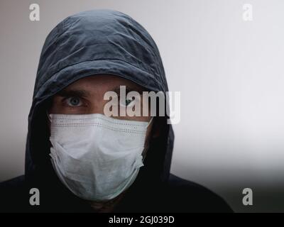 Un homme vêtu de noir avec un capot au-dessus de sa tête et un masque médical blanc. Seuls ses yeux sont visibles. Espace pour le texte. Concept de protection Covid Banque D'Images