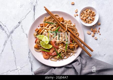 Vue de dessus de l'assiette avec nouilles udon asiatiques, crevettes épicées, tofu frit, arachides, citron vert et oignon vert sur fond de marbre blanc Banque D'Images