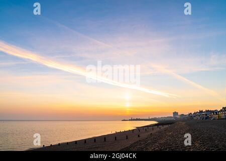 Le soleil se lève dans un ciel orange sur la silhouette de la jetée au loin à la station balnéaire de Kent de Herne Bay. Au premier plan, la plage de galets et les groynes, les briseurs de vagues en bois et une rangée de cabanes de plage qui attrapent la première lumière. Banque D'Images