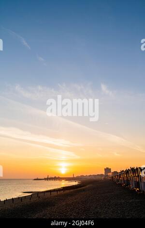 Le soleil se lève dans un ciel orange sur la silhouette de la jetée au loin à la station balnéaire de Kent de Herne Bay. Au premier plan, la plage de galets et les groynes, les briseurs de vagues en bois et une rangée de cabanes de plage qui attrapent la première lumière. Banque D'Images