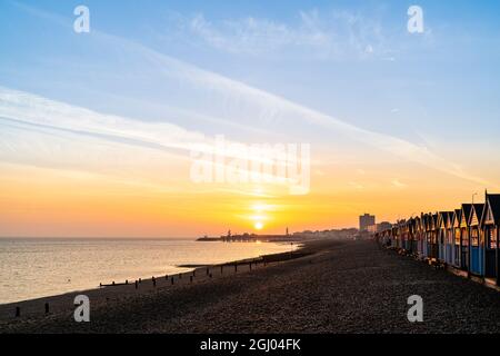 Le soleil se lève dans un ciel orange sur la silhouette de la jetée au loin à la station balnéaire de Kent de Herne Bay. Au premier plan, la plage de galets et les groynes, les briseurs de vagues en bois et une rangée de cabanes de plage qui attrapent la première lumière. Banque D'Images