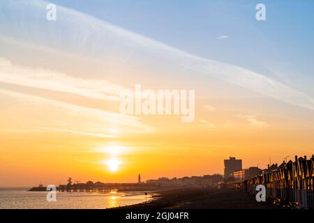 Le soleil se lève dans un ciel orange sur la silhouette de la jetée au loin à la station balnéaire de Kent de Herne Bay. Au premier plan, la plage de galets et les groynes, les briseurs de vagues en bois et une rangée de cabanes de plage qui attrapent la première lumière. Banque D'Images